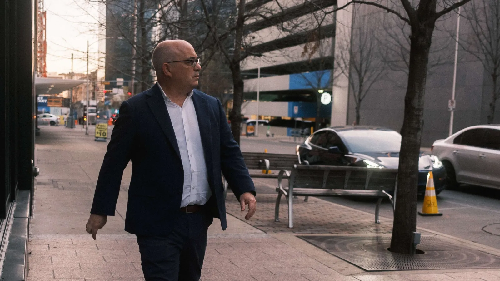 Man with a white shirt and dark blue suit looking to his left, walking towards the camera on a sidewalk in downtown Austin.