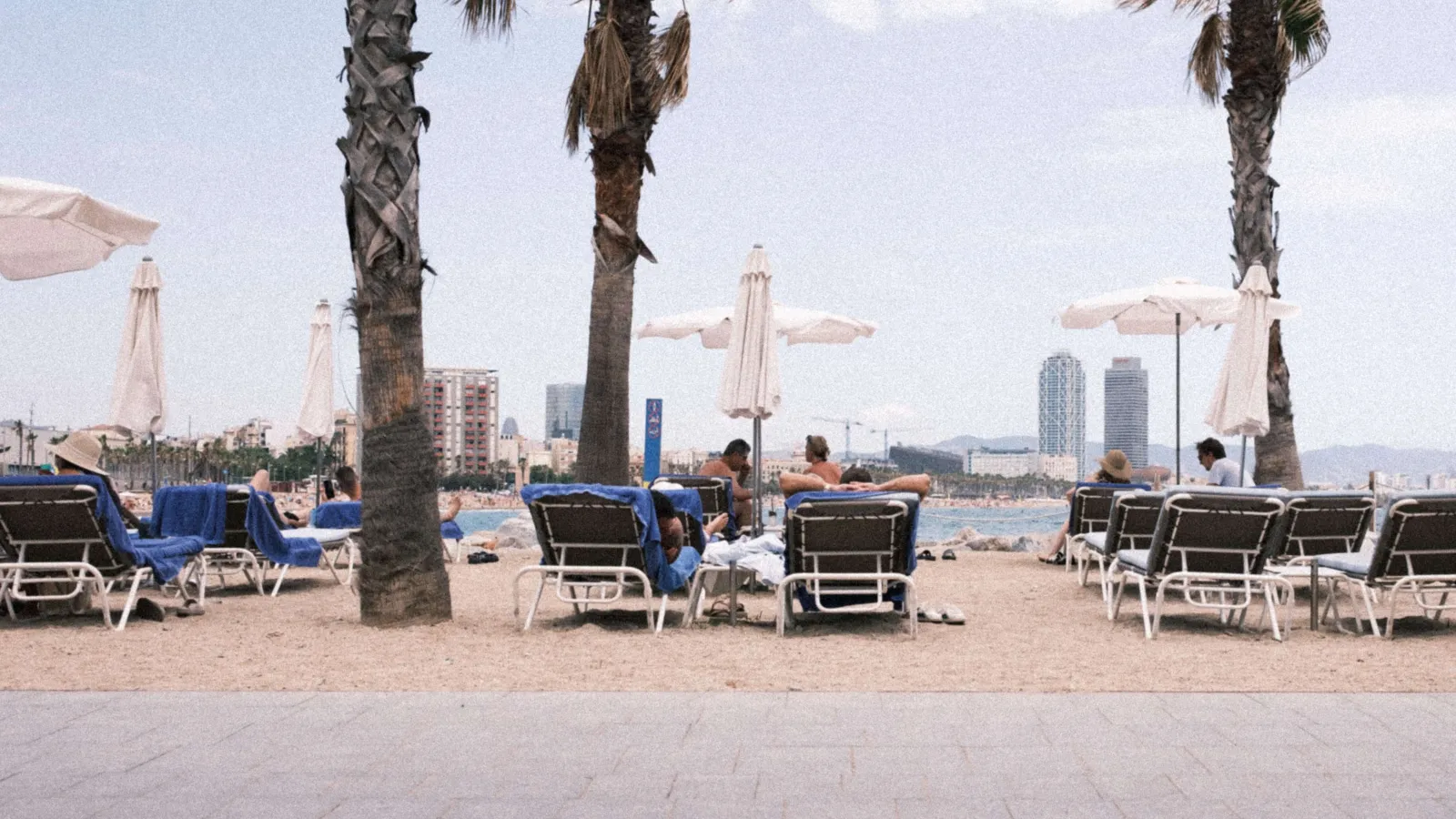 Picture at Barceloneta showing people lying on blue loungers with white sunshades around them at the beach. The ocean is partly visible in the background.