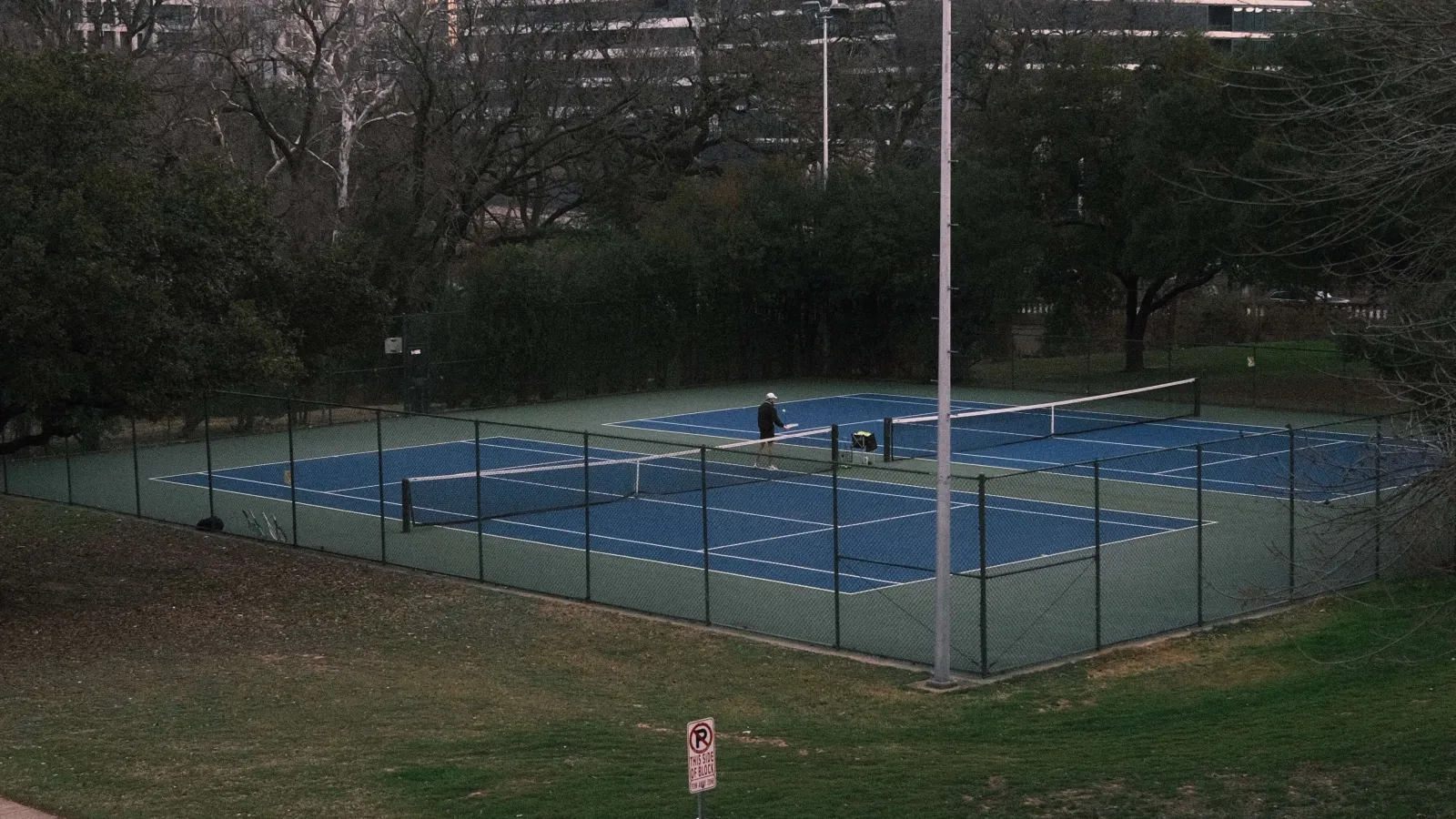 An aerial view of a tennis court surrounded by green grass and trees. A person is seen practicing on the court, with modern buildings in the background.