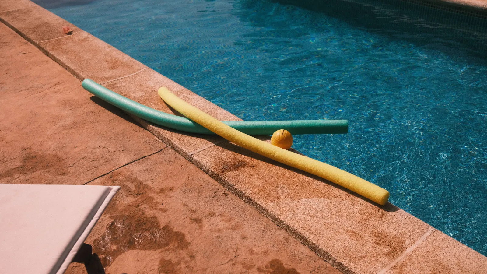 Photo of pool accessories like aqua noodles and a yellow ball lying by the pool.