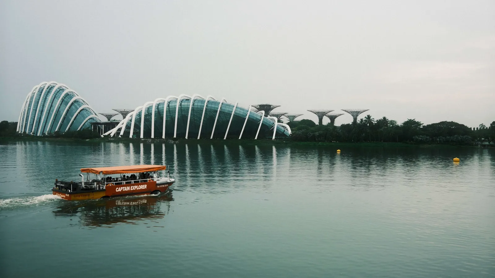 Picture of Gardens by the Bay in Singapore with Marina Bay in between, showing a small red boat with tourists passing by.