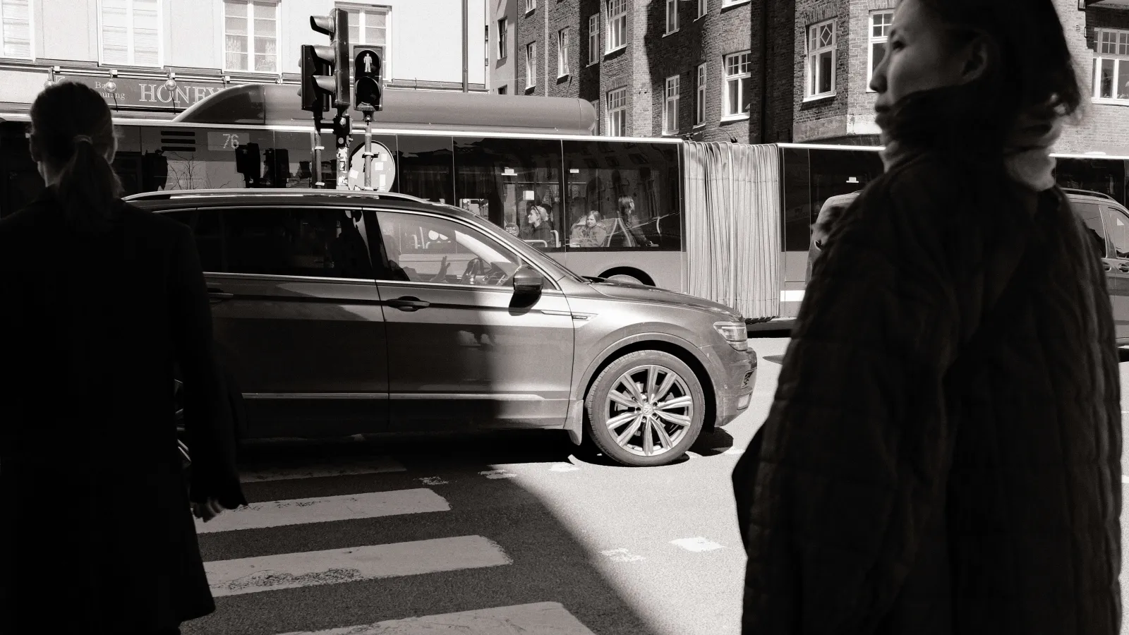 Black-and-white photo at a crosswalk. One person is standing with their back in front of the camera to the right, one person on the left. A car is crossing the street from the left, a bus from the right.